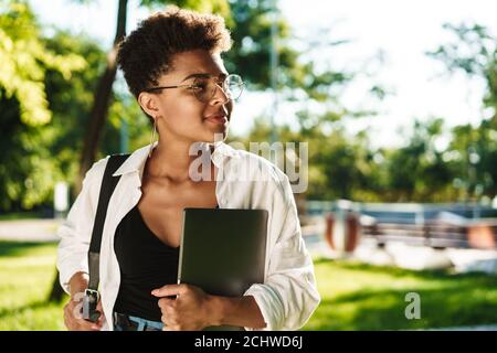 Photo d'une femme africaine heureuse et positive marchant à l'extérieur dans le parc tout en tenant l'ordinateur portable Banque D'Images