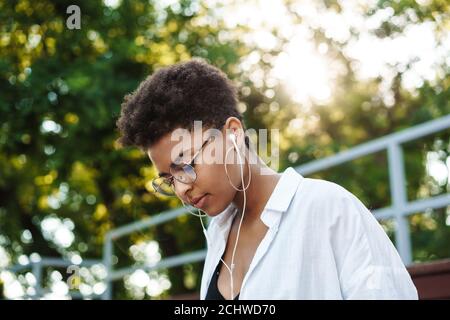 Photo d'une femme africaine sérieuse assise à l'extérieur dans le parc pendant écouter de la musique Banque D'Images