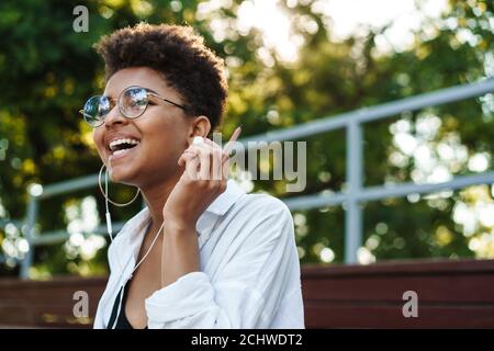 Photo d'une femme africaine optimiste en riant, assise à l'extérieur dans le parc tout en écoutant de la musique Banque D'Images