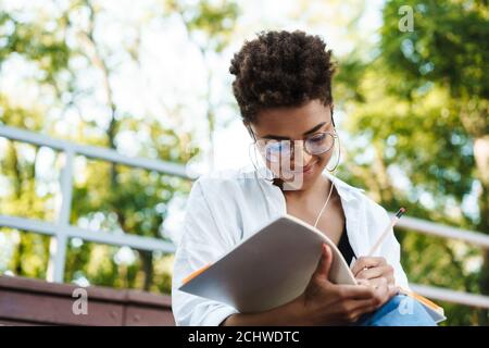 Photo d'une femme africaine concentrée positive assise à l'extérieur dans le parc tout en écoutant de la musique et en lisant des livres Banque D'Images