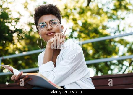 Photo d'une femme africaine concentrée positive assise à l'extérieur dans le parc tout en écoutant de la musique et en lisant des livres Banque D'Images