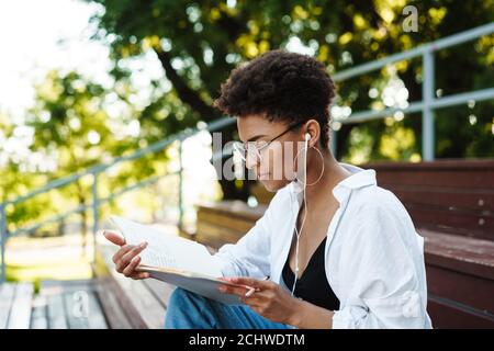 Photo d'une femme africaine concentrée positive assise à l'extérieur dans le parc tout en écoutant de la musique et en lisant des livres Banque D'Images