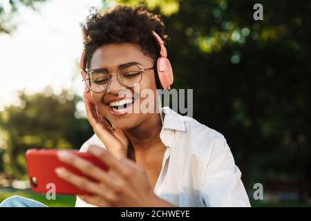 Photo d'une femme africaine en train de rire assise à l'extérieur dans le parc un gazon tout en écoutant de la musique avec des écouteurs et en regardant des vidéos par téléphone portable Banque D'Images