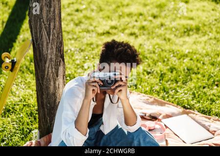 Photo d'une magnifique femme africaine assise à l'extérieur dans le parc sur une pelouse et en tenant la caméra Banque D'Images