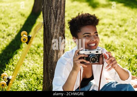 Photo d'une femme africaine souriante et magnifique assise à l'extérieur garez-vous sur une pelouse et tenez l'appareil photo Banque D'Images