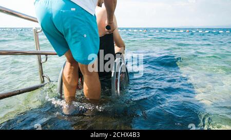 Homme handicapé à la plage nageant sur un fauteuil roulant avec aide sur une rampe d'accès. Banque D'Images