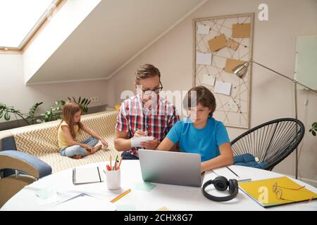 Papa aidant son fils avec des devoirs d'école, garçon apprenant en ligne tandis que sa petite sœur assis sur le canapé sur le fond Banque D'Images