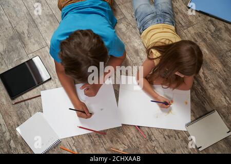 Frères et sœurs jouant ensemble à la maison. Vue de dessus de petit garçon et fille couché sur le plancher en bois et dessin sur des feuilles de papier blanches avec coloré Banque D'Images