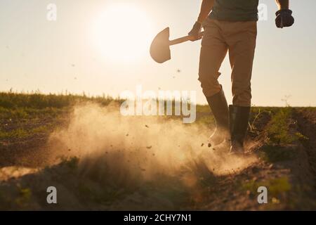 Portrait en coupe basse d'un travailleur masculin méconnaissable qui soufflait de nuages de poussière de bottes en caoutchouc et qui tient la pelle tout en traversant le champ de plantation à la lumière du coucher du soleil, dans l'espace de copie Banque D'Images