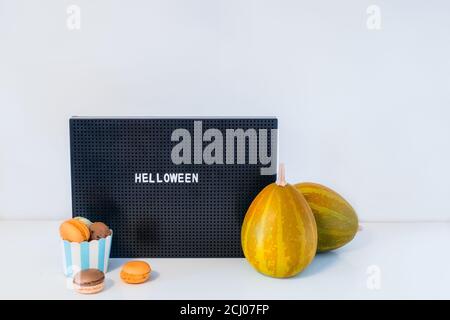 Décor minimaliste pour Halloween - panneau de lettres avec message Helloween, citrouilles décoratives et tasse avec macarons doux sur fond blanc. Décoration festive Banque D'Images