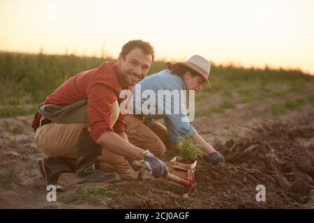 Portrait complet de deux personnes travaillant dans le champ à la plantation de légumes, l'accent sur les jeunes hommes plantant des jeunes arbres en premier plan et souriant à la caméra, l'espace de copie Banque D'Images