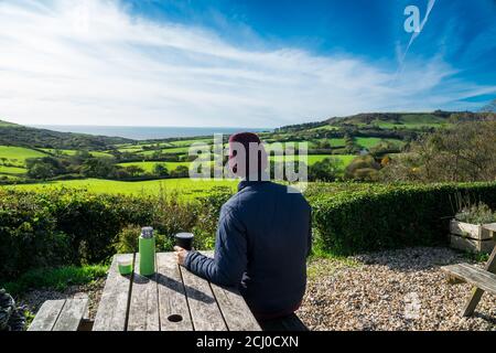 Backview jeune homme dans des vêtements chauds assis dans un café en bois fermier et appréciant la vue sur la nature locale avec thermos de thé sur la table. Vacances d'automne. Nouvelle normale Banque D'Images