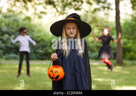 Portrait d'une adolescente souriante habillée comme sorcière posant à l'extérieur et tenant le seau d'Halloween avec les enfants jouant en arrière-plan, espace de copie Banque D'Images