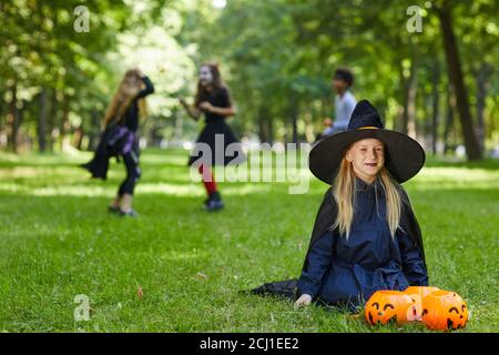 Portrait d'une adolescente souriante habillée comme sorcière pour Halloween assise sur l'herbe verte à l'extérieur avec les enfants jouant en arrière-plan, espace de copie Banque D'Images