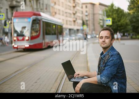 Un homme se trouve près de la ligne de tramway. Un passager attend un tramway assis sur le trottoir, dangereusement près de la voie ferrée avec un ordinateur portable Banque D'Images