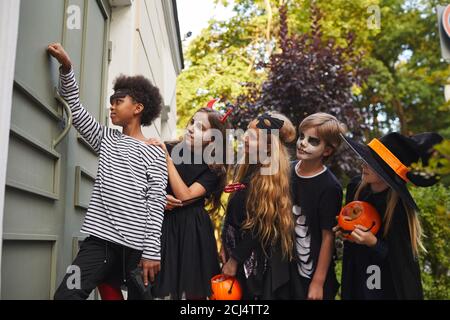 Vue latérale d'un groupe multiethnique d'enfants portant des costumes d'Halloween sonnant la sonnette de porte tout en trick ou en se faisant dorer ensemble, espace de copie Banque D'Images