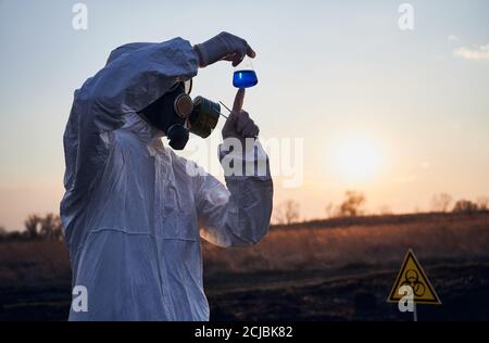 Écologiste mâle en combinaison de protection, masque à gaz et gants tenant un tube à essai avec un liquide bleu tout en étudiant des échantillons de sol sur un territoire brûlé avec un signe de danger biologique. Concept d'écologie et de recherche Banque D'Images