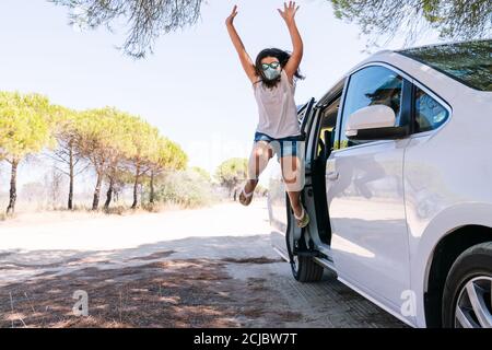 Fille saute de la porte de voiture avec un visage masque et lunettes de soleil brillantes heureux avec ses bras ouverts pour ayant pu profiter des vacances d'été Banque D'Images