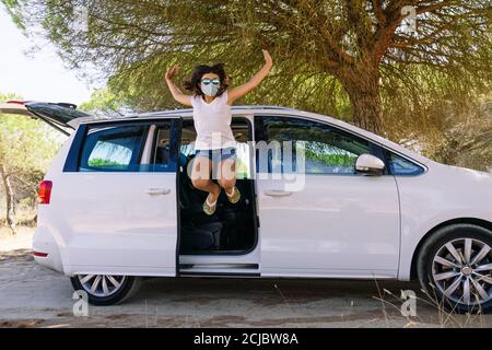 Fille saute de la porte de voiture avec un visage masque et lunettes de soleil brillantes heureux avec ses bras ouverts pour ayant pu profiter des vacances d'été Banque D'Images