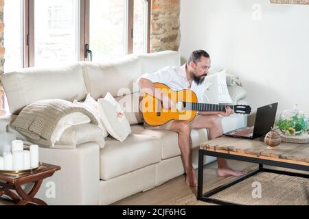 un homme hippster barbu prend des leçons de guitare en ligne sur le canapé à la maison Banque D'Images