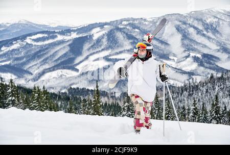 Photo horizontale d'un jeune skieur en costume coloré marchant le long d'une colline enneigée, cardant ses skis, contre des montagnes étonnantes en arrière-plan. Vue de face, espace de copie, pleine longueur Banque D'Images