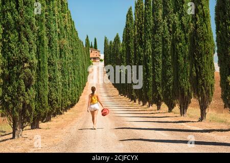 Vue arrière d'une femme marchant sur une route de terre bordée de cyprès toscans par une journée ensoleillée. Villa italienne à l'horizon avec fond bleu ciel. Banque D'Images