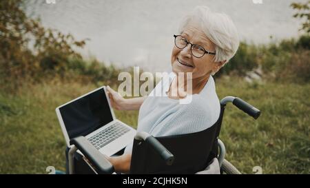 Femme âgée heureuse en fauteuil roulant utilisant un ordinateur portable et souriant vers l'appareil photo. Photo de haute qualité Banque D'Images
