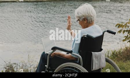 Une femme adulte handicapée agite à la caméra de l'ordinateur portable pendant l'appel vidéo près de la rivière. Photo de haute qualité Banque D'Images