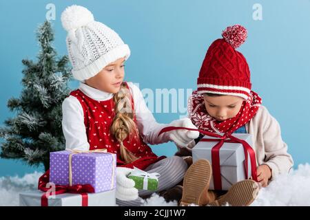 enfants en tenue d'hiver assis sur la neige près de firs et tenue présente isolée sur bleu Banque D'Images