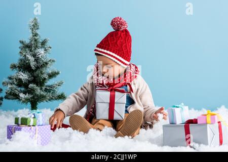 garçon en chapeau et tenue d'hiver assis près de cadeaux et arbre de noël tout en touchant la neige isolée sur bleu Banque D'Images