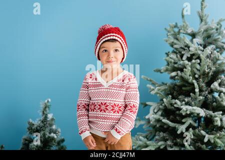 garçon en tenue d'hiver debout près des arbres de noël sur bleu Banque D'Images