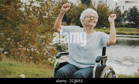 Une femme âgée dansant avec les mains dans le fauteuil roulant près de la rivière le jour de l'automne. Photo de haute qualité Banque D'Images