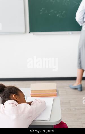 attention sélective à une écolière afro-américaine fatiguée qui dormait à son bureau pendant que l'enseignant se tient près du tableau de surveillance Banque D'Images