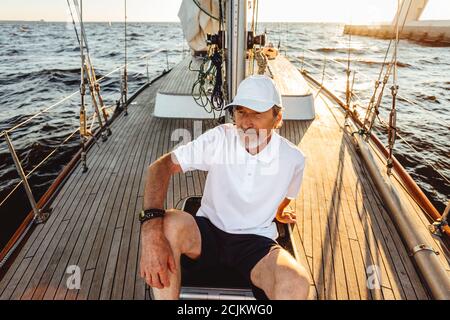 Homme senior assis sur un pont de son yacht et profitant du coucher du soleil. Capitaine mature se détendant sur un voilier. Banque D'Images