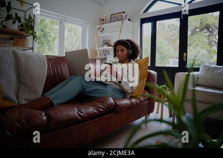 Jeune femme afro-américaine se détendant sur un canapé à l'écoute de musique tout en effectuant un paiement en ligne à l'aide d'une carte bancaire et d'un ordinateur portable Banque D'Images