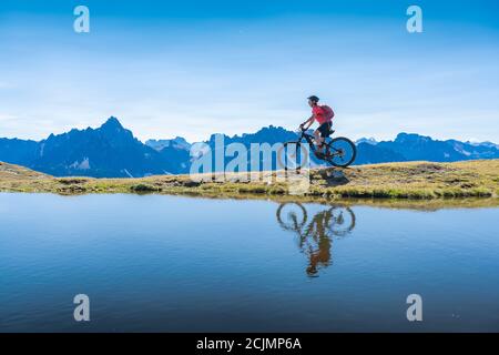 Belle femme à cheval sur son vélo électrique de montagne les trois pics Dolomites, se reflétant dans l'eau bleue d'un lac de montagne froid Banque D'Images