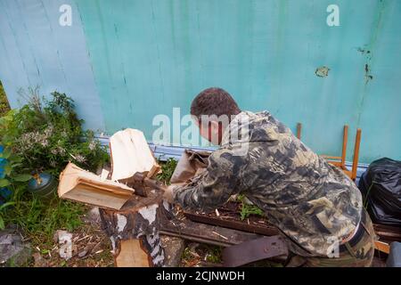 Un homme dans un costume de camouflage hache le bois avec un hache avec une poignée en bois et une lame en fer le village ou à la datcha à côté d'un métal clôture pour kindl Banque D'Images