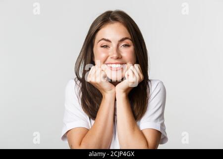 Gros plan d'une petite fille de brunette souriante et charmante, penchée sur les mains et regardant avec admiration, fond blanc Banque D'Images