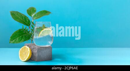 Un verre d'eau avec citron et menthe sur le piédestal dans un style tendance sur fond bleu. Bannière Banque D'Images