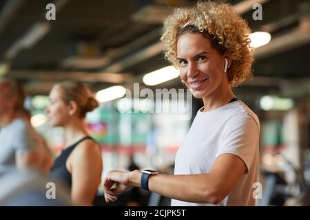 Portrait d'une femme mûre avec des cheveux bouclés souriant à l'appareil photo tout en vérifiant son pouls sur la montre-bracelet pendant l'entraînement Banque D'Images