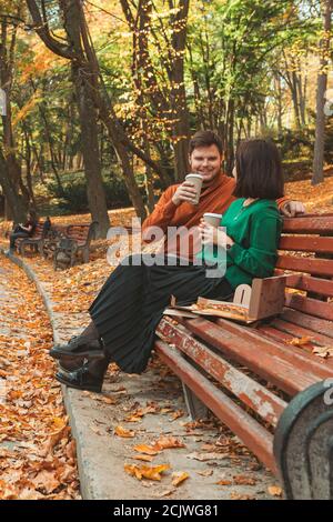 un jeune joli couple assis sur le banc du parc buvant du café manger de la pizza en parlant l'un l'autre Banque D'Images