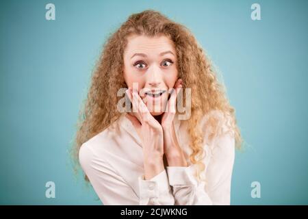 Émerveillement belle blonde jeune femme avec les mains sur les joues isolées sur fond bleu Banque D'Images