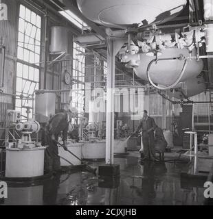 Années 1950, les travailleurs industriels historiques lavant un plancher d'usine, l'un pulvérisant de l'eau, l'autre utilisant une mop, Angleterre, Royaume-Uni. Banque D'Images