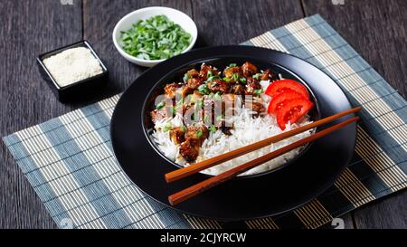 Recette à base de plantes de tofu teriyaki avec riz arrosé de scallion, graines de sésame servies avec du riz, tomates tranchées, sur un bol noir avec baguettes Banque D'Images