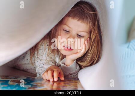 Belle petite fille lisant le livre couché sur le lit sous la couverture dans la chambre. Banque D'Images