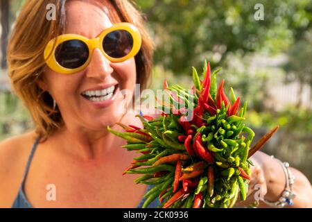 Gros plan sur un paquet de piments verts et rouges frais tenir par une belle femme de brunette caucasienne riante avec des lunettes de soleil jaunes élégantes. Banque D'Images