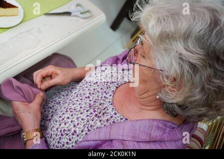 Vue depuis le dessus de la grand-mère avec des cheveux gris, couture à la main à la table de cuisine et un gâteau. Elle porte des lunettes et une robe à fleurs violettes Banque D'Images