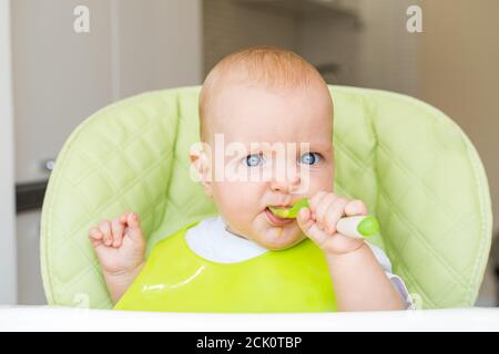 un petit enfant aux yeux bleus et aux cheveux blonds est assis dans une chaise haute et mange le premier appât. Une alimentation saine. Bébé 6 mois. Banque D'Images