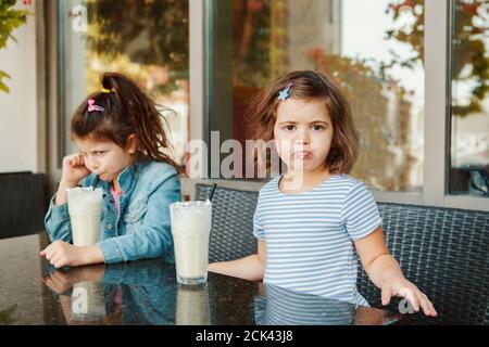 Deux petites sœurs caucasiennes dreuses drôles boivent des milk-shakes dans un café. Amis filles se querelles. Desserts d'été froids pour les enfants. Joyeux authentique Banque D'Images