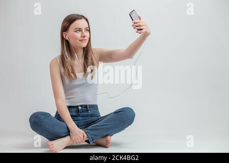 Jeune femme souriante à cheveux justes faisant des photos de selfie sur smartphone sur fond blanc. Espace de copie. Pleine longueur Banque D'Images